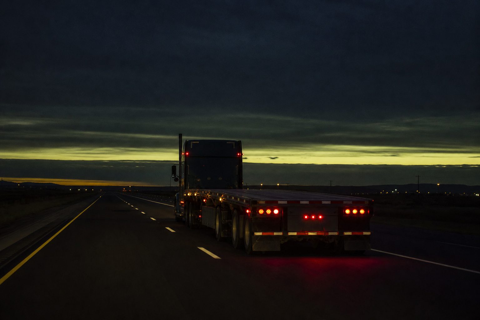 Empty flatbed trailer running a dark interstate at dusk with amber marker lights and dashed lane lines